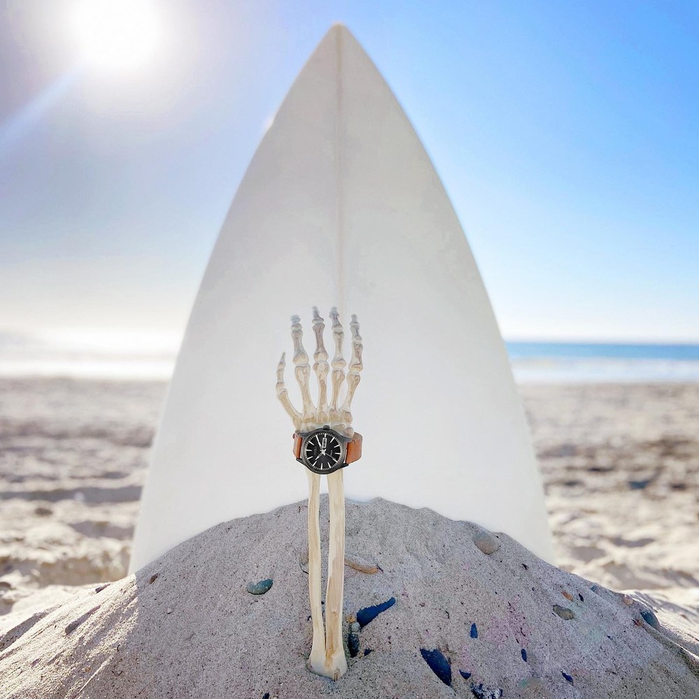 Planche de surf plantee dans le sable, montre Nixon posee dessus