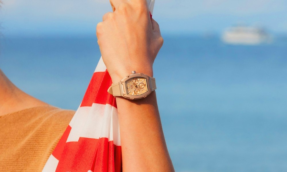 Bras portant une montre etanche doree devant la mer, parasol raye rouge et blanc