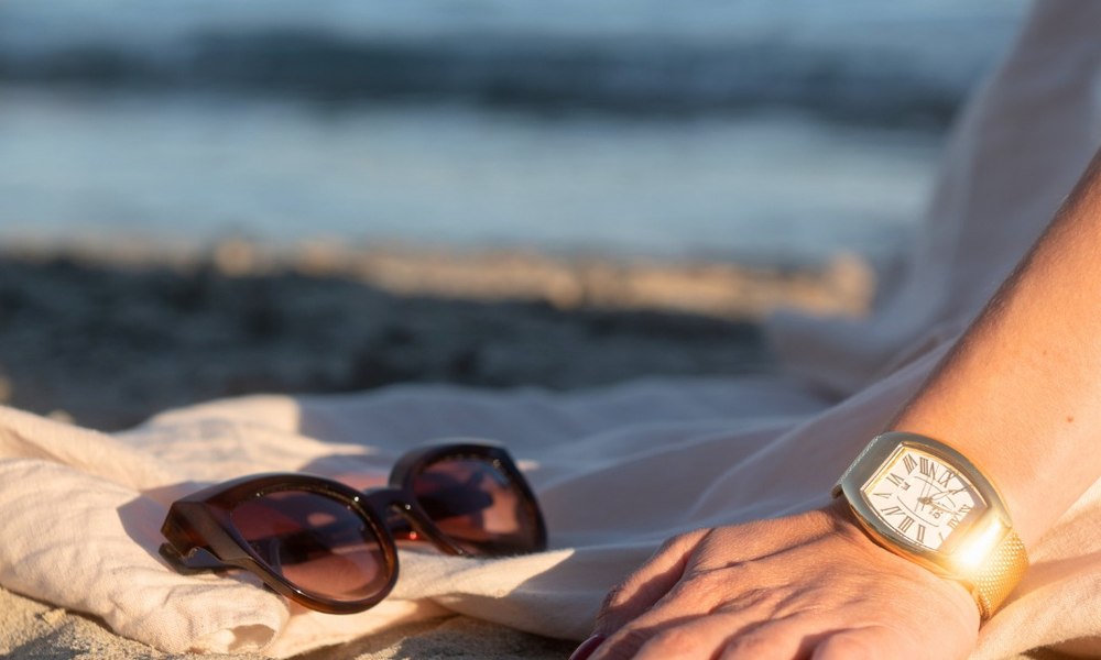 Mains posees sur le sable chaud pres de lunettes de soleil, montre doree au poignet