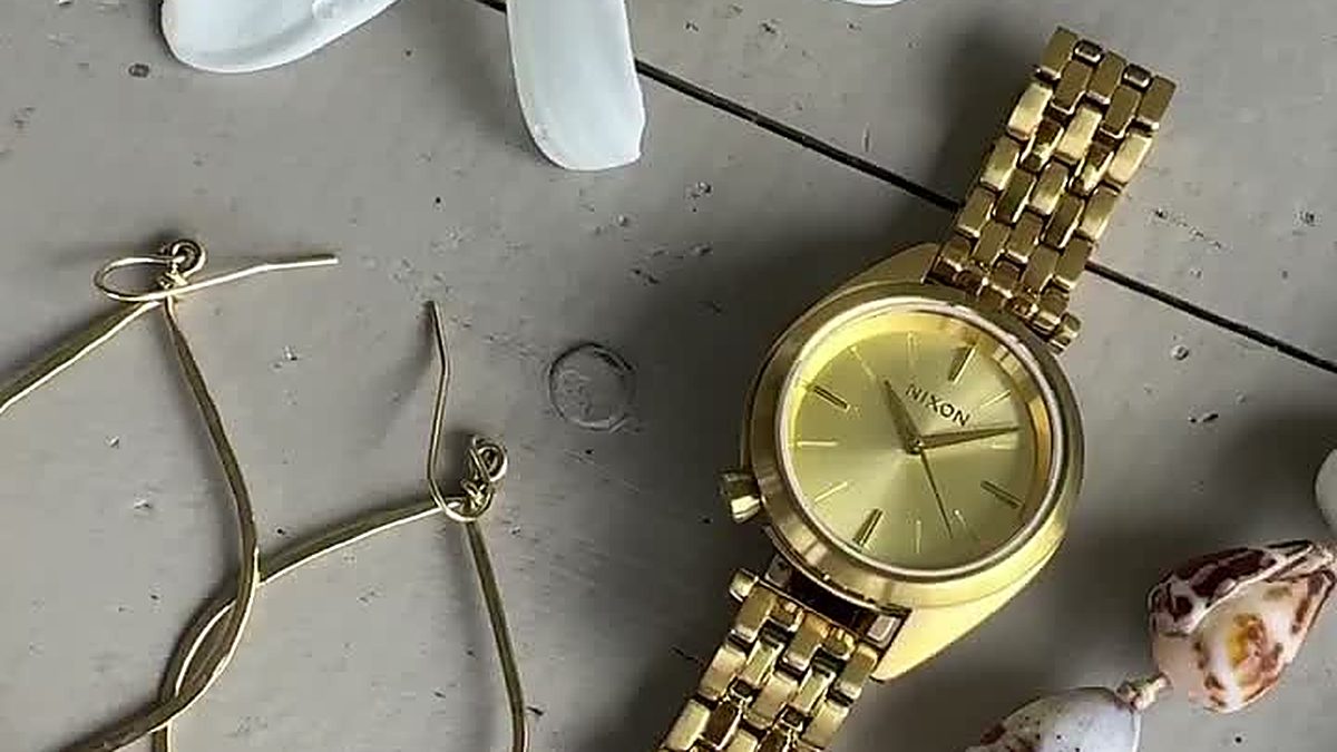 Montre dorée Nixon Time Teller posée à plat entre une fleur de tiaré, des boucles d'oreilles dorées et un collier de coquillages, composition estivale lumière dorée, guide de la montre dorée 2026 Club de la Montre