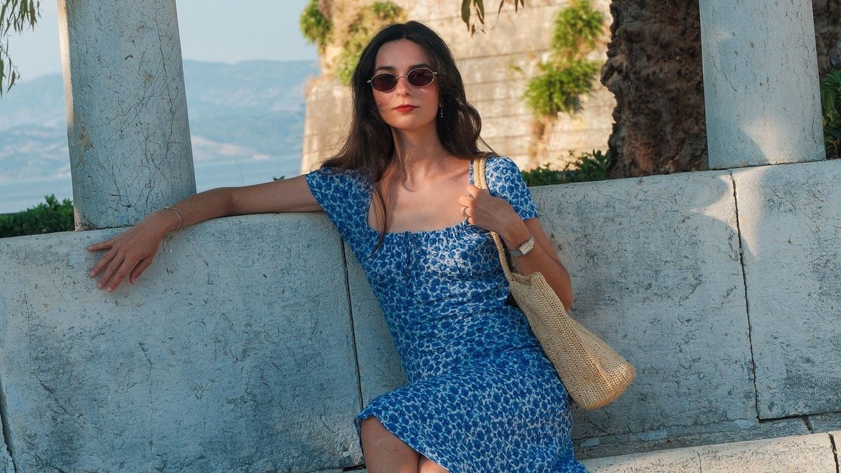 A woman in a blue summer dress wearing a French watch, sitting facing the sea
