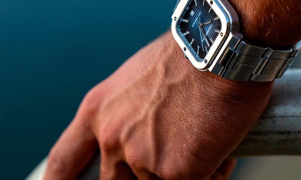 Poignet masculin portant une montre a boitier carre en acier devant la mer